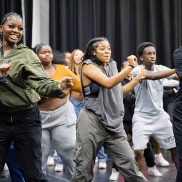 A photo of a group of young people in a rehearsal space warming up by dancing.
