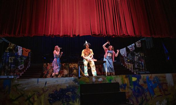 One ADP 2024 Company member sits wearing a party hat in the middle of the stage whilst two people sing either side of him and the stage curtain falls.