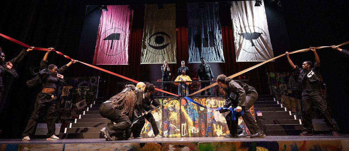 The ADP 2024 Company performing on stage, with several cast members pulling long bits of thick ribbon in different directions on the stage.