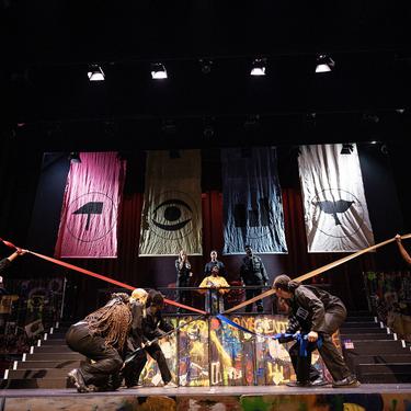 The ADP 2024 Company performing on stage, with several cast members pulling long bits of thick ribbon in different directions on the stage.