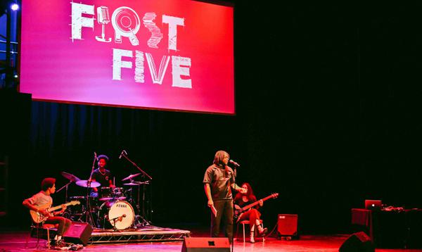A photo of Yomi Sode on stage at the start of First Five with the House band behind him and a projector screen saying First Five.