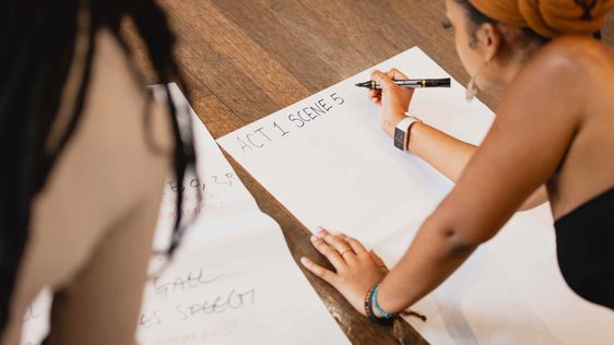 A photo of large bits of white paper on the floor and two young individuals writing script ideas on it.