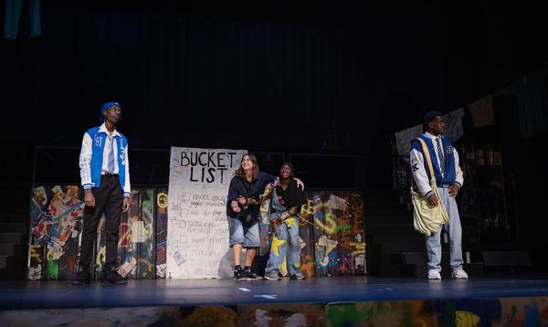 Four ADP 2024 Company members are acting together on stage, with a giant Bucket List sign behind them.