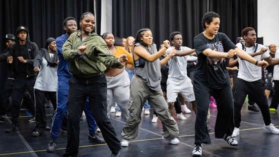 A photo of a group of young people in a rehearsal space warming up by dancing.