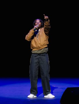 A young person is performing on the ,main stage holding the mic close to their mouth as they sign into the microphone and the other hand is in the air pointing to the audience.