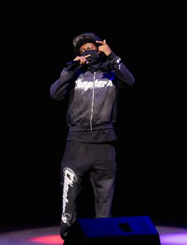 A headline act is wearing a dark tracksuit and cap and is preforming on stage, holding a microphone and gesturing with their other hand. The background is black with colored stage lights on the floor.