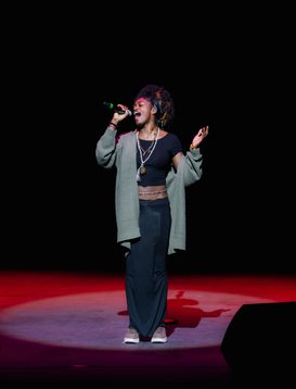 A person wearing a dark tracksuit and cap performs on stage, holding a microphone and gesturing with their other hand. The background is black with colored stage lights on the floor.