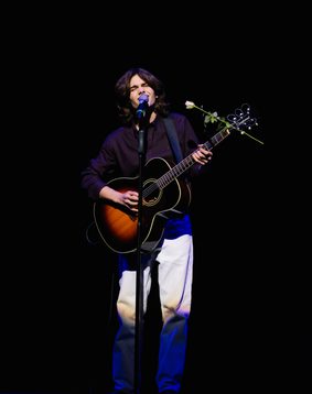 A young person wearing a deep purple shirt and white trousers is playing the guitar on Hackney Empire's main stage and they also have a red rose attached to the guitar.
