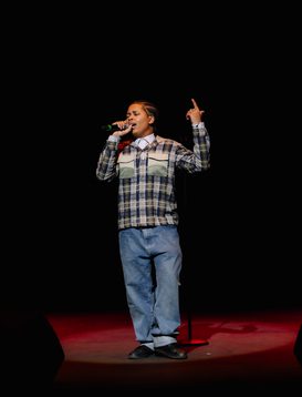 A young person wearing a a brown checked shift and blue jeans performing live on on Hackney Empire's main stage singing into the microphone