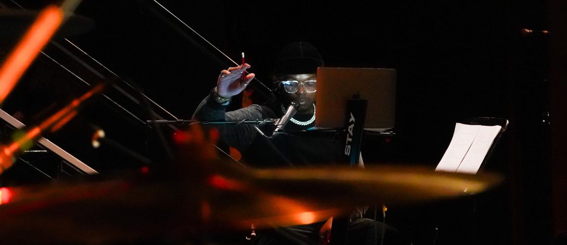 A musician sits at a workstation with a laptop and microphone in a dimly lit rehearsal or recording space. Wearing a black beanie, glasses, and a chain necklace, they raise one hand while looking toward the laptop. Out-of-focus drums and warm orange stage lights create a layered foreground, with sheet music visible on a stand to the side.