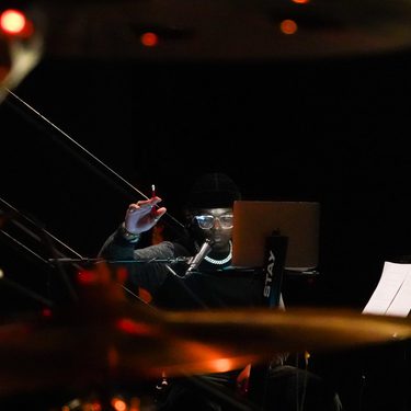 A musician sits at a workstation with a laptop and microphone in a dimly lit rehearsal or recording space. Wearing a black beanie, glasses, and a chain necklace, they raise one hand while looking toward the laptop. Out-of-focus drums and warm orange stage lights create a layered foreground, with sheet music visible on a stand to the side.