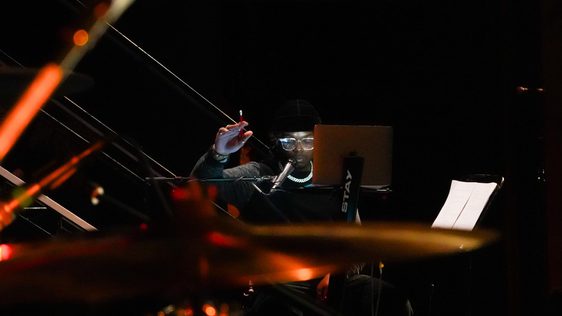 A musician sits at a workstation with a laptop and microphone in a dimly lit rehearsal or recording space. Wearing a black beanie, glasses, and a chain necklace, they raise one hand while looking toward the laptop. Out-of-focus drums and warm orange stage lights create a layered foreground, with sheet music visible on a stand to the side.