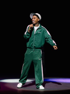 Headliner Kilo Keyz on stage wearing a green tracksuit and white hat, is holding the microphone to his mouth to perform.